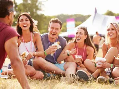 young men and women friends sitting on the grass while eating and laughing at a music festival